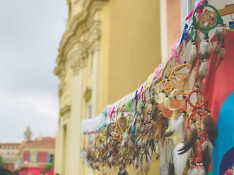 A collection of colorful dreamcatchers hangs in an outdoor market setting. The dreamcatchers feature feathers, beads, and intricate web patterns against the backdrop of historic architecture. The building in the background is softly focused, adding depth to the scene.