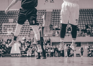Two basketball players are captured in mid-air on a court, wearing jerseys with numbers, while an audience watches in the background. The image has a vintage sepia tone with blurred spectators.