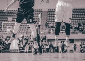 Two basketball players are captured in mid-air on a court, wearing jerseys with numbers, while an audience watches in the background. The image has a vintage sepia tone with blurred spectators.