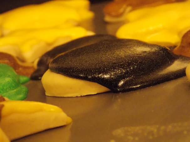 A close-up of cookies topped with colorful icing, including black, yellow, green, and brown shades. The icing has a shiny texture, and the cookies are resting on a flat surface.