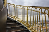 A spiral staircase with golden railings overlooking a cityscape. The sky is overcast with grey clouds, and the city buildings are visible in the background. The railings have intricate patterns and appear well-maintained, adding an elegant touch to the scene.