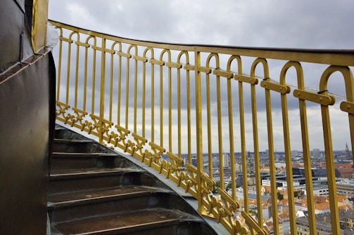 A spiral staircase with golden railings overlooking a cityscape. The sky is overcast with grey clouds, and the city buildings are visible in the background. The railings have intricate patterns and appear well-maintained, adding an elegant touch to the scene.