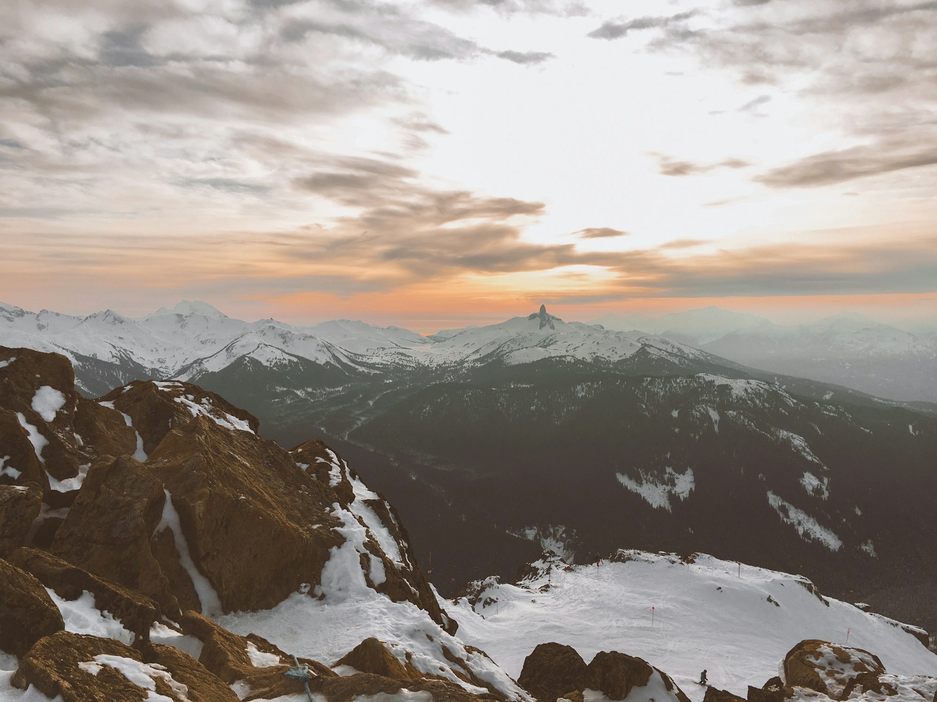 A breathtaking panoramic view from a snowy mountaintop showing endless forested valleys and jagged peaks illuminated by the soft light of a winter sunset.
