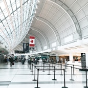 A spacious airport terminal with a high arched ceiling, featuring a large Canadian flag. The terminal is equipped with check-in counters, digital screens, and black crowd control stanchions lining walkways. There are people walking and a few individuals standing near the counters, likely travelers checking in.