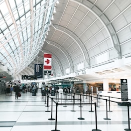 A spacious airport terminal with a high arched ceiling, featuring a large Canadian flag. The terminal is equipped with check-in counters, digital screens, and black crowd control stanchions lining walkways. There are people walking and a few individuals standing near the counters, likely travelers checking in.