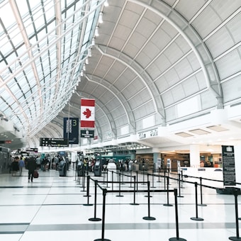 A spacious airport terminal with a high arched ceiling, featuring a large Canadian flag. The terminal is equipped with check-in counters, digital screens, and black crowd control stanchions lining walkways. There are people walking and a few individuals standing near the counters, likely travelers checking in.