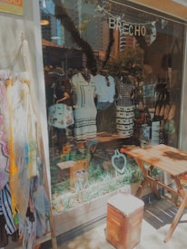 A shop window display featuring various garments, including dresses and shorts, arranged on mannequins. The storefront is decorated with plants at the base, and a wooden table and seat are placed outside. A selection of scarves is draped on a stand nearby, and a small blackboard sign with writing is visible. The background shows reflections of urban buildings.