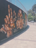 A vibrant street mural featuring large, realistic paintings of hands and faces adorned with butterflies. The artwork is situated on a curved wall beside a sidewalk. The background is predominantly black, which makes the warm colors of the paintings stand out. Cars and trees are visible in the street scene nearby.