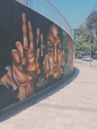 A vibrant street mural featuring large, realistic paintings of hands and faces adorned with butterflies. The artwork is situated on a curved wall beside a sidewalk. The background is predominantly black, which makes the warm colors of the paintings stand out. Cars and trees are visible in the street scene nearby.