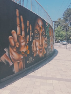 A vibrant street mural featuring large, realistic paintings of hands and faces adorned with butterflies. The artwork is situated on a curved wall beside a sidewalk. The background is predominantly black, which makes the warm colors of the paintings stand out. Cars and trees are visible in the street scene nearby.