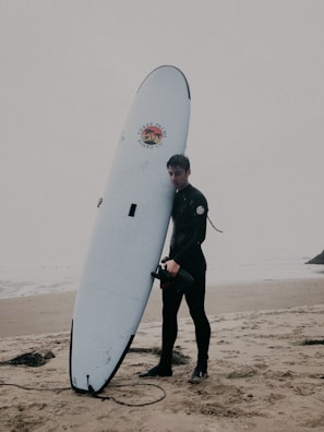 A person wearing a wetsuit is standing on a sandy beach holding a large surfboard upright. The background is overcast, giving a moody and calm atmosphere. The surfboard has a logo with the words 'Three Palms Board Co.' and a palm tree design. The ocean and a faint view of the beach stretch out behind them.
