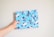 A hand is holding a wrapped gift covered in light blue wrapping paper adorned with cartoon penguin illustrations wearing winter hats and scarves. The gift is neatly tied with a matching bow, and the background is a plain off-white wall.