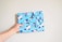A hand is holding a wrapped gift covered in light blue wrapping paper adorned with cartoon penguin illustrations wearing winter hats and scarves. The gift is neatly tied with a matching bow, and the background is a plain off-white wall.