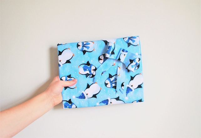 A hand is holding a wrapped gift covered in light blue wrapping paper adorned with cartoon penguin illustrations wearing winter hats and scarves. The gift is neatly tied with a matching bow, and the background is a plain off-white wall.
