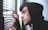 A person in a black hoodie is peering through the slats of a set of window blinds, placing their fingers gently on the blinds to hold them apart. The individual appears to be focused intently outside, possibly observing something of interest or concern.