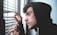 A person in a black hoodie is peering through the slats of a set of window blinds, placing their fingers gently on the blinds to hold them apart. The individual appears to be focused intently outside, possibly observing something of interest or concern.