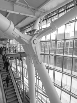 A large structural beam is prominently featured, intricately designed and bolted in an architectural space. The environment includes large glass windows allowing natural light to flood into the space. Several people are using an escalator, ascending or descending, adding a sense of movement to the scene.