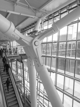 A large structural beam is prominently featured, intricately designed and bolted in an architectural space. The environment includes large glass windows allowing natural light to flood into the space. Several people are using an escalator, ascending or descending, adding a sense of movement to the scene.