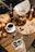 A cozy coffee setup features a Chemex coffee maker with freshly brewed coffee, next to a copper coffee pot. A white cup filled with coffee is placed on a saucer, accompanied by a spoon. There are scattered coffee beans and cherries on the table, along with sliced dried apricots. Two Polaroid photos are also on display, beside a loaf of bread and a vase with decorative branches.