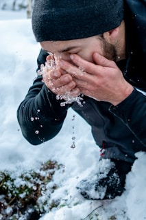 A person wearing a black beanie and dark clothing leans forward to splash water onto their face from a snow-covered surface. The water droplets are captured mid-air as the person holds their hands close to their face.