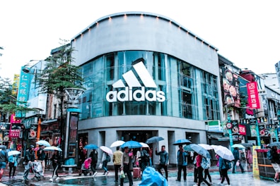 A busy urban street scene features a modern store with a large glass facade displaying the 'Adidas' logo. Numerous pedestrians walk on the wet pavement, many carrying umbrellas, suggesting rainy weather. Bright signage in various languages is visible on the surrounding buildings, adding a vibrant urban feel.