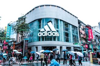 A busy urban street scene features a modern store with a large glass facade displaying the 'Adidas' logo. Numerous pedestrians walk on the wet pavement, many carrying umbrellas, suggesting rainy weather. Bright signage in various languages is visible on the surrounding buildings, adding a vibrant urban feel.