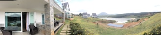 A panoramic view of a serene lakeside area with modern houses. The scene includes a patio with chairs and tables, a stone pathway, green grass, small plants, several modern houses, a lake, and a distant mountain range under a cloudy sky.