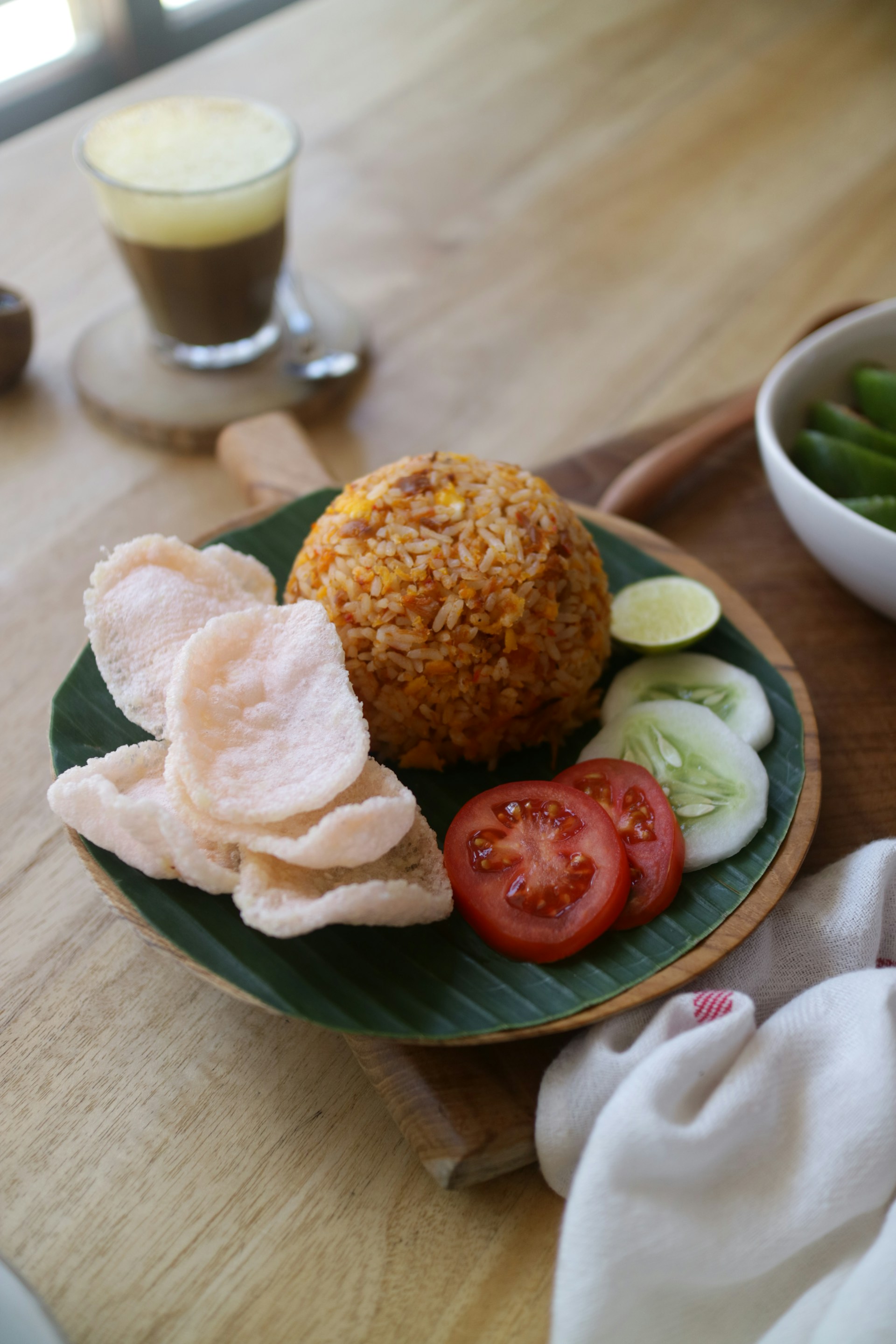 A dish consisting of a serving of fried rice topped with a few prawn crackers, served with sliced tomatoes and cucumbers on a banana leaf. A lime wedge is placed next to the rice. In the background, there is a glass cup with a beverage, possibly coffee or tea, on a coaster. The setting is on a wooden table with a portion of a white cloth visible.