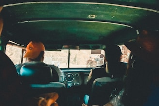 The interior view of a vehicle with several people inside. The image is taken from the backseat, looking towards the front where two individuals are seated, one wearing an orange cap. The atmosphere is dimly lit, with a warm and earthy color palette visible through the vehicle's windows.