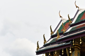 An ornate temple roof is adorned with intricate golden decorations. The roof features traditional sloping edges with green and red tiles, highlighted by stylized design elements pointing upwards. The backdrop is a cloudy sky, emphasizing the roof's detailed craftsmanship.