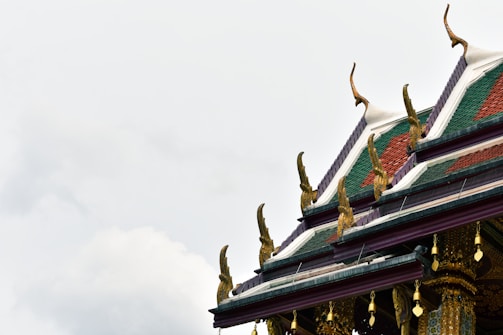 An ornate temple roof is adorned with intricate golden decorations. The roof features traditional sloping edges with green and red tiles, highlighted by stylized design elements pointing upwards. The backdrop is a cloudy sky, emphasizing the roof's detailed craftsmanship.
