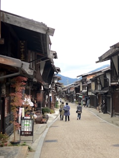 A traditional Japanese street with wooden buildings lining both sides. Three people walk down the road, which is flanked by small shops and decorative plants. Mountains are visible in the background under a cloudy sky.