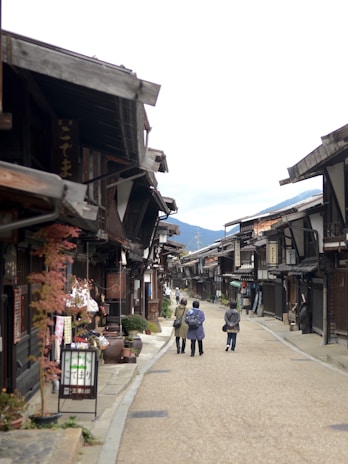A traditional Japanese street with wooden buildings lining both sides. Three people walk down the road, which is flanked by small shops and decorative plants. Mountains are visible in the background under a cloudy sky.