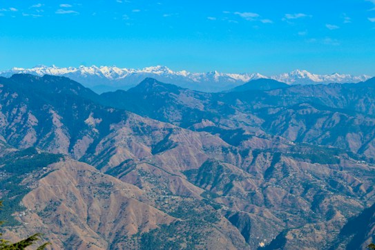 Expansive mountain landscape featuring rugged peaks and ridges, with snow-capped mountains in the background. The image suggests a natural, remote setting with clear blue skies above.