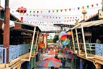 A colorful marketplace with vibrant triangular bunting hanging overhead. Various shops and stalls are visible on both sides, and a large circular, tropical-themed sign hangs in the middle. Multi-colored umbrellas are open on the ground level, adding to the festive atmosphere. People are walking and browsing around the area.