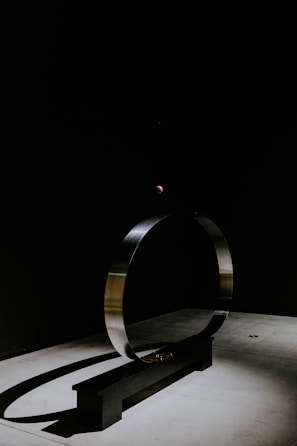 A minimalist metal sculpture featuring a large circular ring stands on a rectangular base. The artwork is set against a dark background with a small, illuminated sphere at the top, casting a subtle shadow on the floor.
