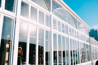 A glass building with large window panes reflecting sunlight. The angular perspective shows a decorative arch with stained glass elements near the top. Inside, chairs and tables are visible. The bright sky and a hint of urban skyline can be seen in the background.