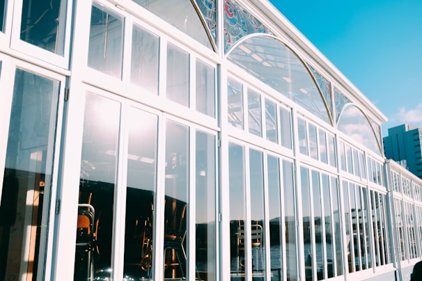 A glass building with large window panes reflecting sunlight. The angular perspective shows a decorative arch with stained glass elements near the top. Inside, chairs and tables are visible. The bright sky and a hint of urban skyline can be seen in the background.