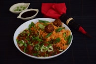 A bowl of tantalizing Indian rice and meatball dish garnished with fresh herbs and green onions. The dish is accompanied by two small bowls, one containing green chilies and the other a dark sauce, placed on a dark surface. Additionally, there is a red cloth napkin with a bamboo-themed chopstick set placed next to the bowl.