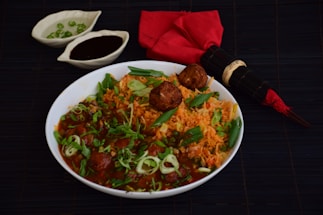 A bowl of tantalizing Indian rice and meatball dish garnished with fresh herbs and green onions. The dish is accompanied by two small bowls, one containing green chilies and the other a dark sauce, placed on a dark surface. Additionally, there is a red cloth napkin with a bamboo-themed chopstick set placed next to the bowl.