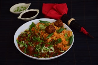 A bowl of tantalizing Indian rice and meatball dish garnished with fresh herbs and green onions. The dish is accompanied by two small bowls, one containing green chilies and the other a dark sauce, placed on a dark surface. Additionally, there is a red cloth napkin with a bamboo-themed chopstick set placed next to the bowl.