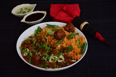 A bowl of tantalizing Indian rice and meatball dish garnished with fresh herbs and green onions. The dish is accompanied by two small bowls, one containing green chilies and the other a dark sauce, placed on a dark surface. Additionally, there is a red cloth napkin with a bamboo-themed chopstick set placed next to the bowl.