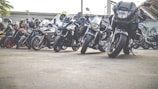 A row of motorcycles parked on the roadside with several people standing nearby. The motorcycles vary in design and color, ranging from sporty models to cruisers, and are aligned closely together. Some riders are wearing helmets, and there's a sense of gathering or meeting in an outdoor setting with a building in the background.