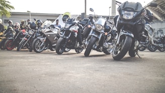 A row of motorcycles parked on the roadside with several people standing nearby. The motorcycles vary in design and color, ranging from sporty models to cruisers, and are aligned closely together. Some riders are wearing helmets, and there's a sense of gathering or meeting in an outdoor setting with a building in the background.