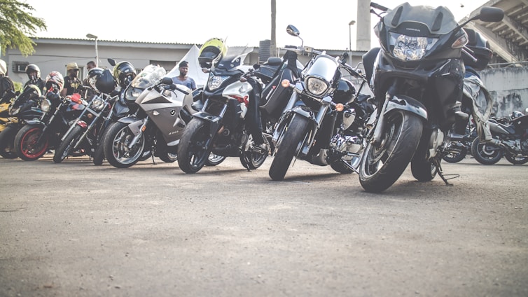 A row of motorcycles parked on the roadside with several people standing nearby. The motorcycles vary in design and color, ranging from sporty models to cruisers, and are aligned closely together. Some riders are wearing helmets, and there's a sense of gathering or meeting in an outdoor setting with a building in the background.