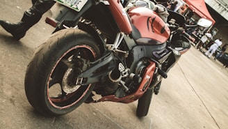 A red and black sport motorcycle is parked on a concrete surface. The motorcycle has a sleek design with intricate details on the body and tires, visible from a side angle. In the background, people are gathered near a red tent and various structures. The scene suggests a casual outdoor event or gathering.