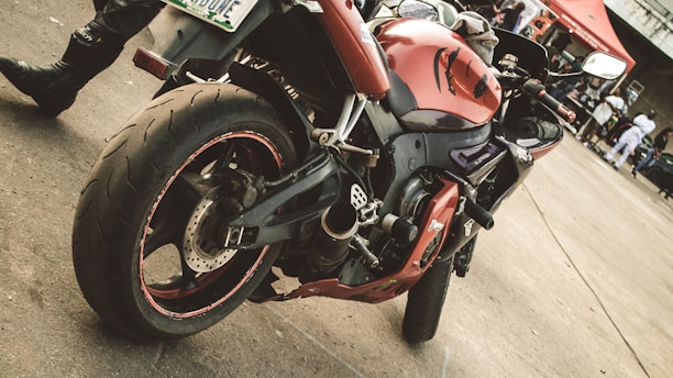 A red and black sport motorcycle is parked on a concrete surface. The motorcycle has a sleek design with intricate details on the body and tires, visible from a side angle. In the background, people are gathered near a red tent and various structures. The scene suggests a casual outdoor event or gathering.