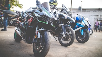 Several high-performance motorcycles are parked together in an outdoor area. The motorcycles are predominantly black with accents of green, blue, and silver. In the background, a few people are standing, and more motorcycles are visible.