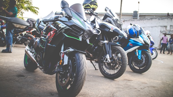 Several high-performance motorcycles are parked together in an outdoor area. The motorcycles are predominantly black with accents of green, blue, and silver. In the background, a few people are standing, and more motorcycles are visible.