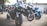 Several high-performance motorcycles are parked together in an outdoor area. The motorcycles are predominantly black with accents of green, blue, and silver. In the background, a few people are standing, and more motorcycles are visible.